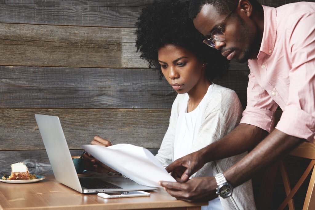 cooperation and team work. african american businessman in pink shirt holding papers, explaining details of a bargain to his beautiful partner with afro haircut while having a meeting at a coffee shop